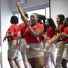 A group of young adults in red polo shirts and khaki shorts performing enthusiastically, with one woman in the foreground holding a microphone and raising her arm, eyes closed and smiling deeply.