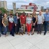 A diverse group of nine adults smiling and posing for a photo on a rooftop patio with a cityscape and overcast sky in the background. Some are wearing attire with a red "M" logo.