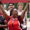A group of cheerful young adults walking on a campus pathway, with a woman in the middle holding a red sign with "LAUREN" written on it.